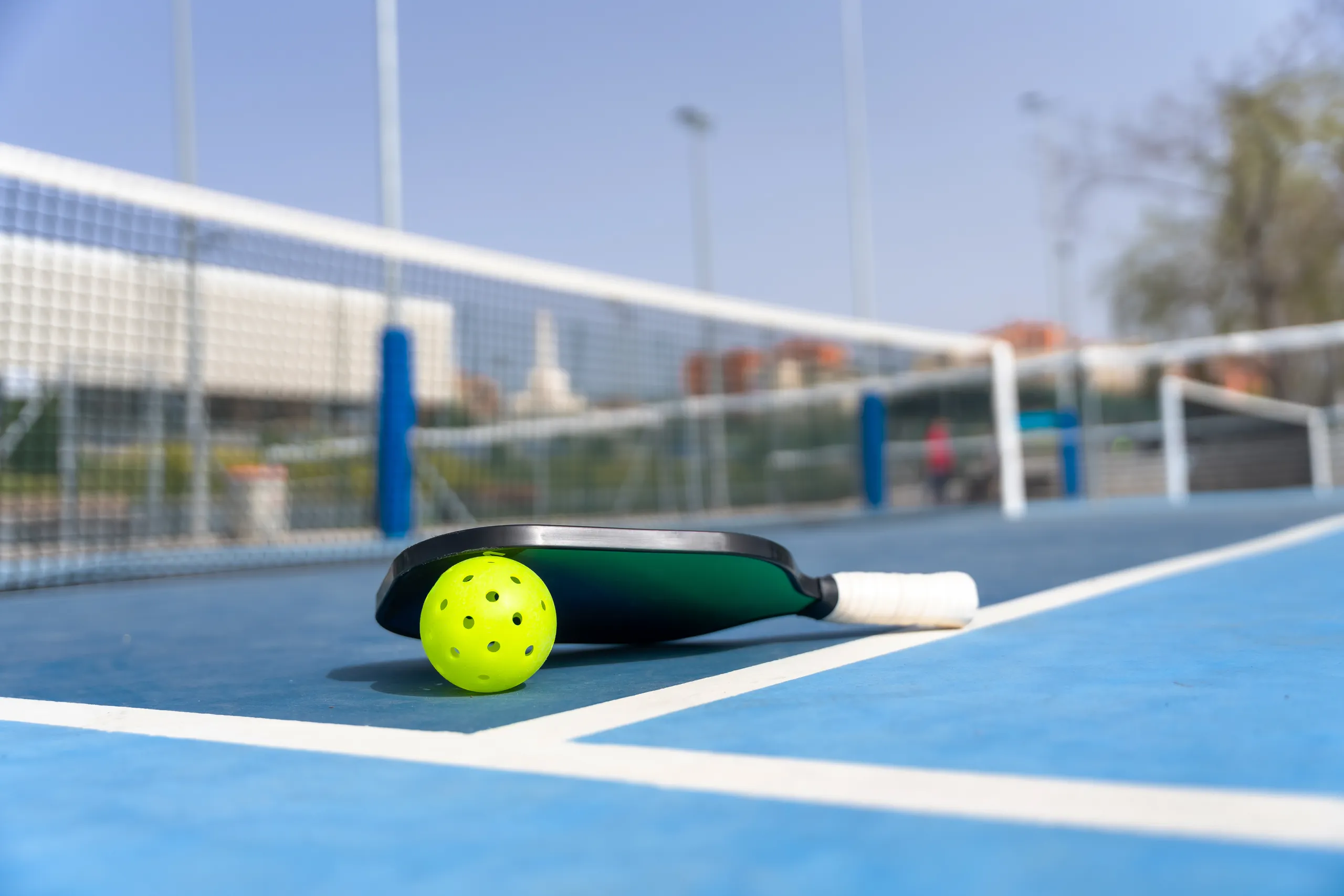 A pickleball paddle and a yellow pickleball ball resting on a blue court with a net in the background.
