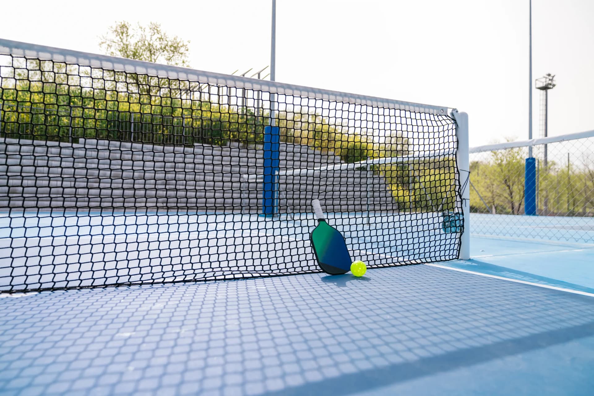 A pickleball paddle and ball resting against the net on a blue outdoor court.