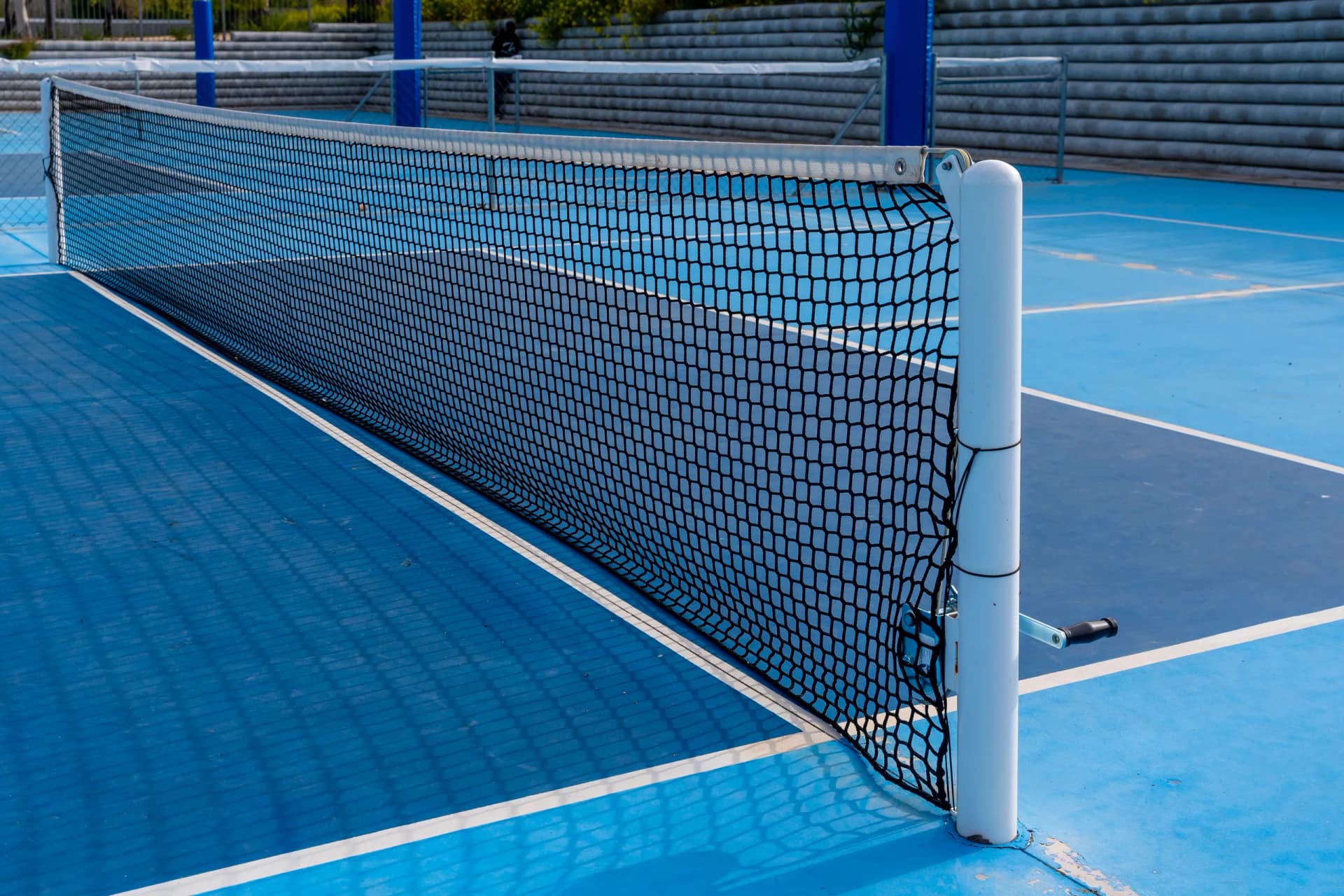 A close-up view of a black net and white post on a blue tennis or pickleball court with white lines.